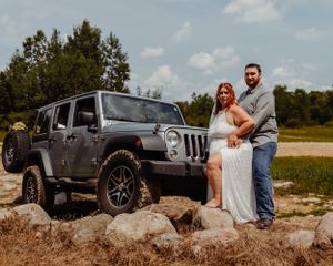 Wedding couple stands on rock formation with their grey Jeep Wrangler parked behind them.