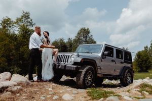 A couple in wedding attire pose together beside a silver Jeep Wrangler on a rocky overlook.