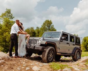 A couple shares an intimate moment next to their silver Jeep Wrangler on rocky terrain.