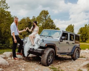 A couple in wedding attire poses against their lifted silver Jeep Wrangler on a rocky landscape.