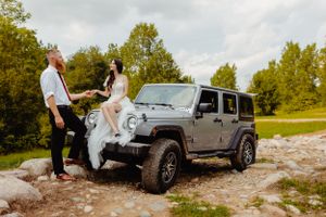 A couple in wedding attire stands beside their silver Jeep Wrangler during an outdoor photo session.
