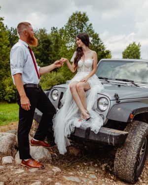 A couple shares a tender moment while posing with their Jeep Wrangler on rocky terrain.