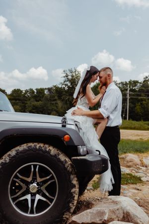 A romantic wedding couple poses on a silver Jeep Wrangler with rugged off-road tires in an outdoor setting.