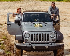 A decorated silver Jeep Wrangler with floral arrangements on its hood parked in a rustic outdoor setting.