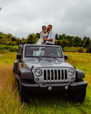 A wedding couple stands through the open roof of their silver Jeep Wrangler in a grassy field.