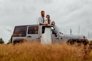A couple in wedding attire stands atop their silver Jeep Wrangler in a golden field at sunset.