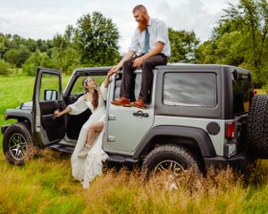A wedding couple poses casually on their silver Jeep Wrangler surrounded by green countryside.