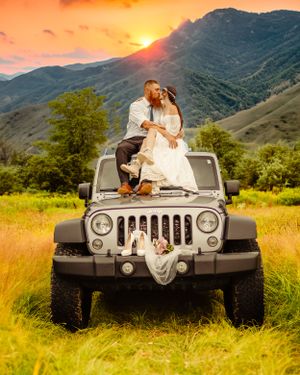 A romantic wedding couple sits atop a silver Jeep Wrangler at sunset in a mountain valley.