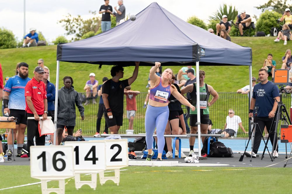 Athletes compete in outdoor track and field event under dark canopy tent.