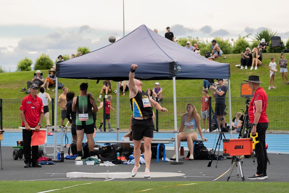 Athletes and officials gather under blue canopy tent at outdoor track competition.