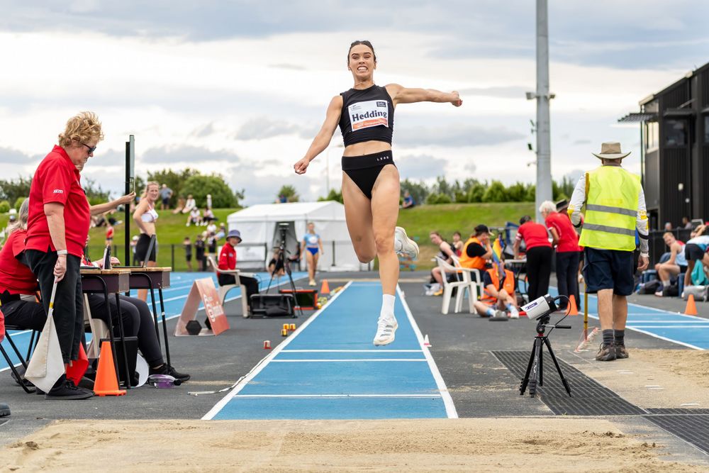Athlete mid-flight during long jump competition with officials and spectators observing.