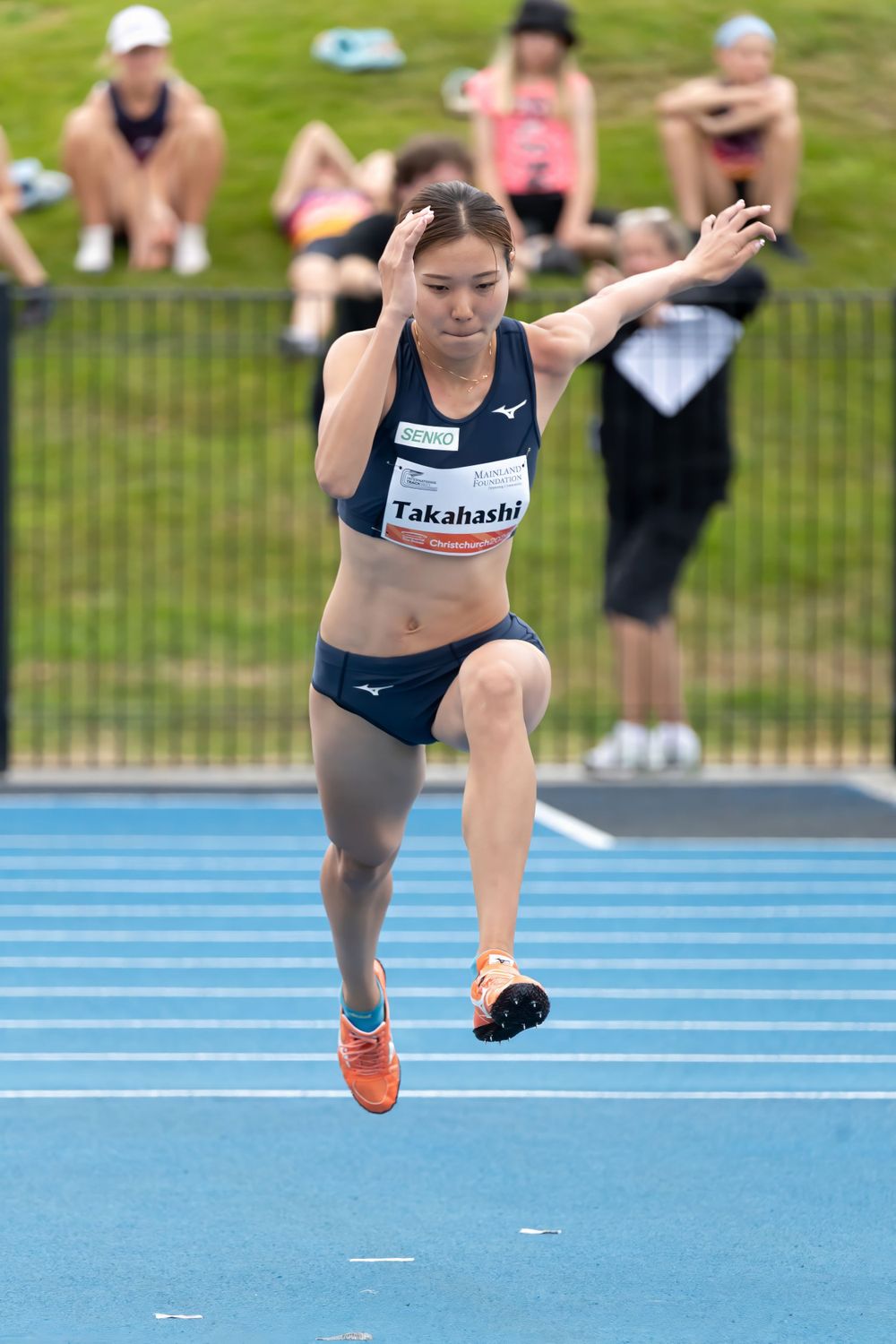 Athlete practicing jumping technique on blue track with spectators in background.