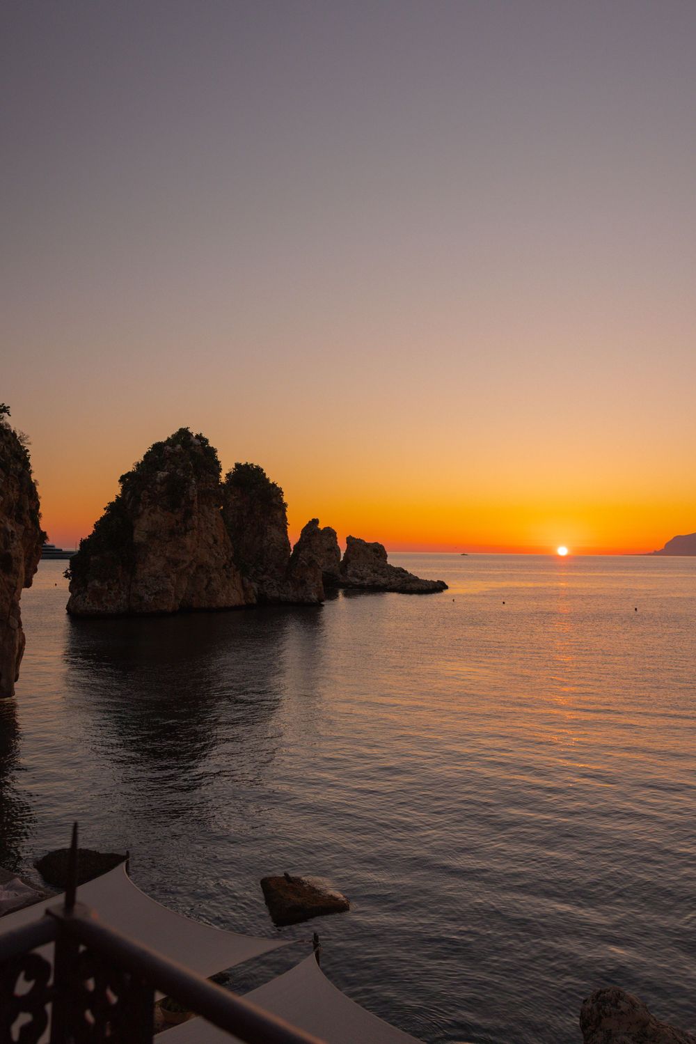 Dramatic sunset silhouettes rocky coastal formations against an orange sky over calm Mediterranean waters.