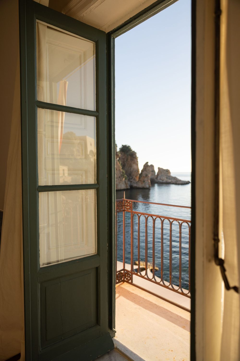 Open balcony door framing view of coastal rock formations and Mediterranean Sea.