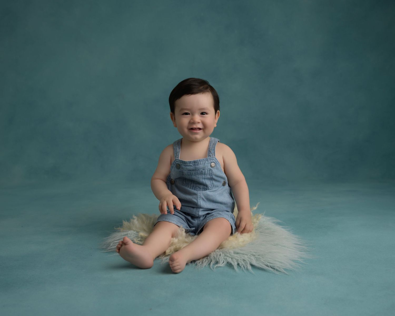 Baby in denim overalls sits on gray fur against teal backdrop during photoshoot.
