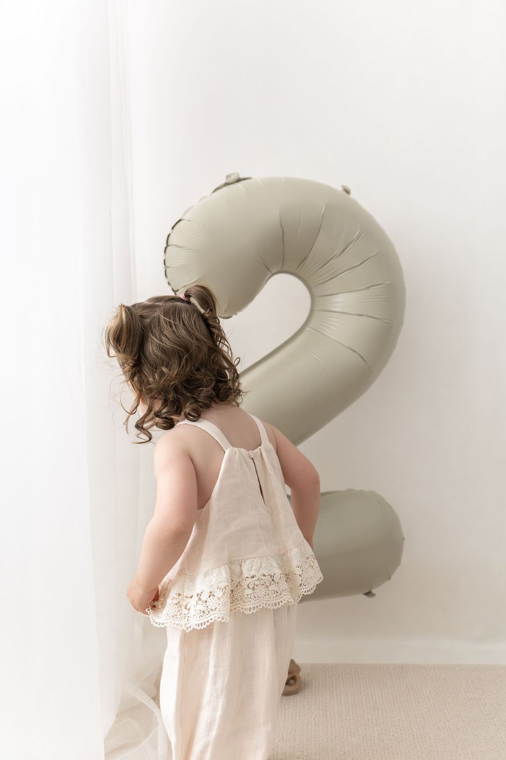 little girl in lace dress standing beside a large sage green number 2 balloon for her second birthday photoshoot Nottingham studio