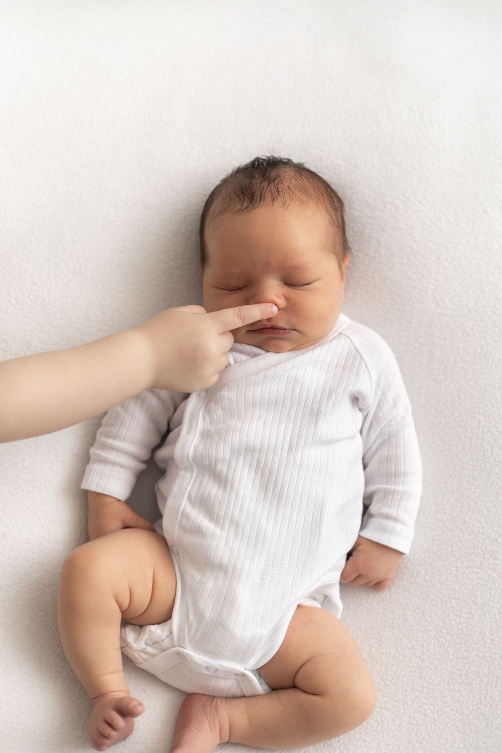 Newborn baby in white onesie lying on white blanket while baby siblings finger gently touches tiny nose Nottingham Newborn photographer