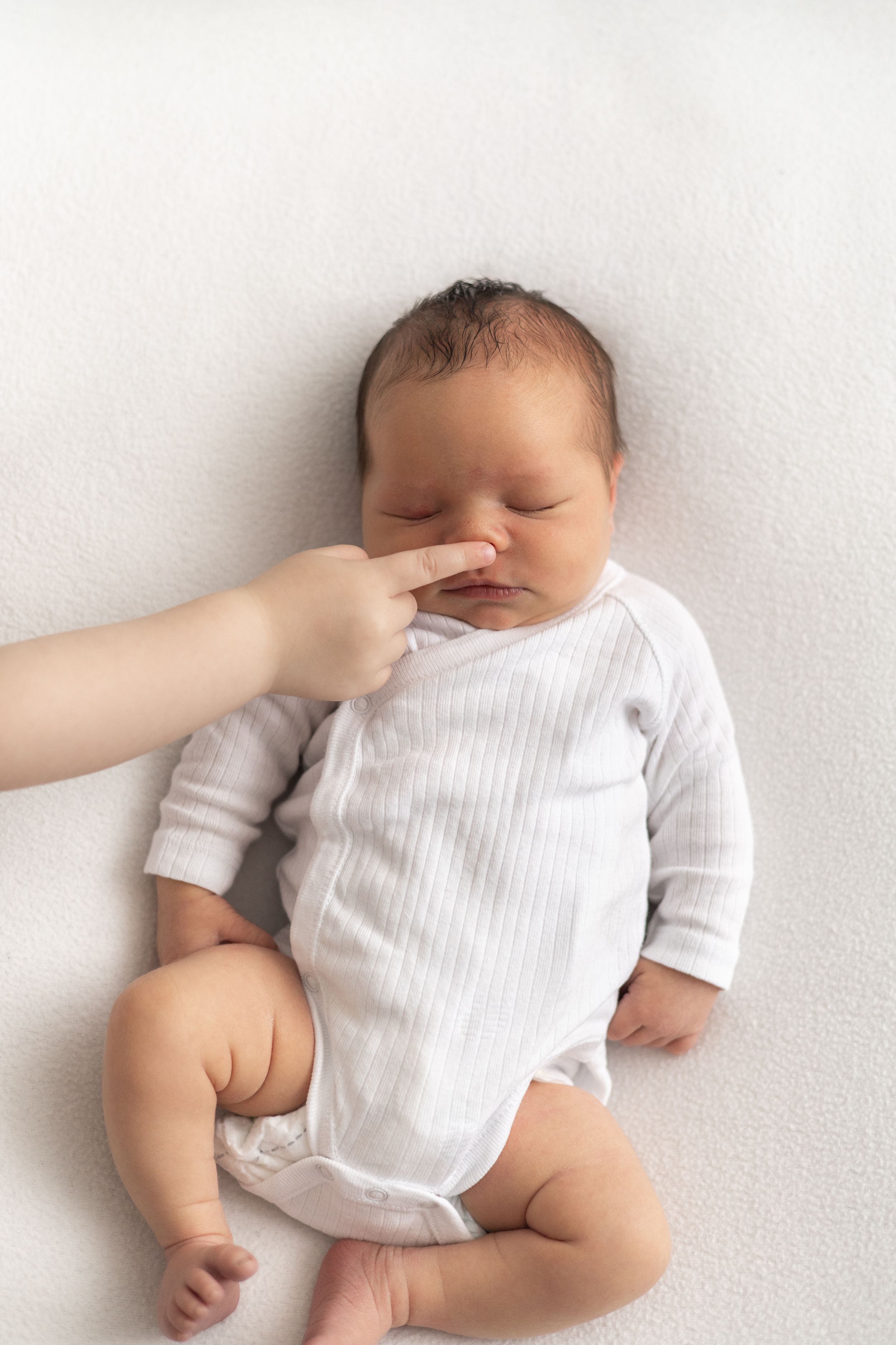 Newborn baby in white onesie lying on white blanket while baby siblings finger gently touches tiny nose Nottingham Newborn photographer