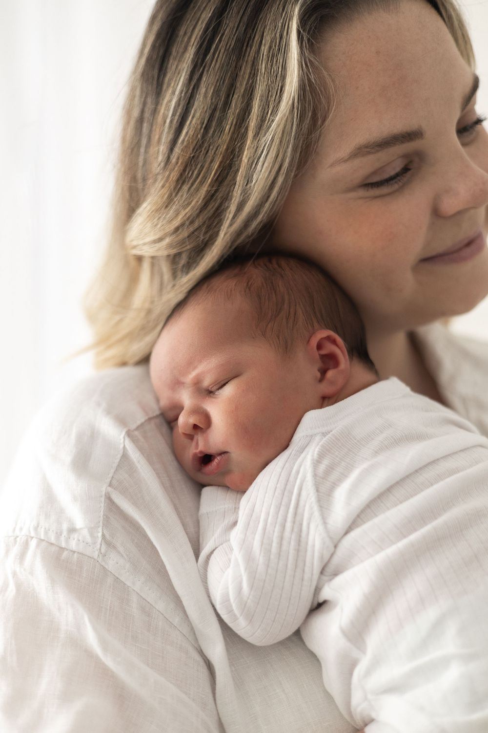A mother tenderly holds her sleeping newborn baby against her chest, both dressed in soft white clothing Nottingham Newborn photographer