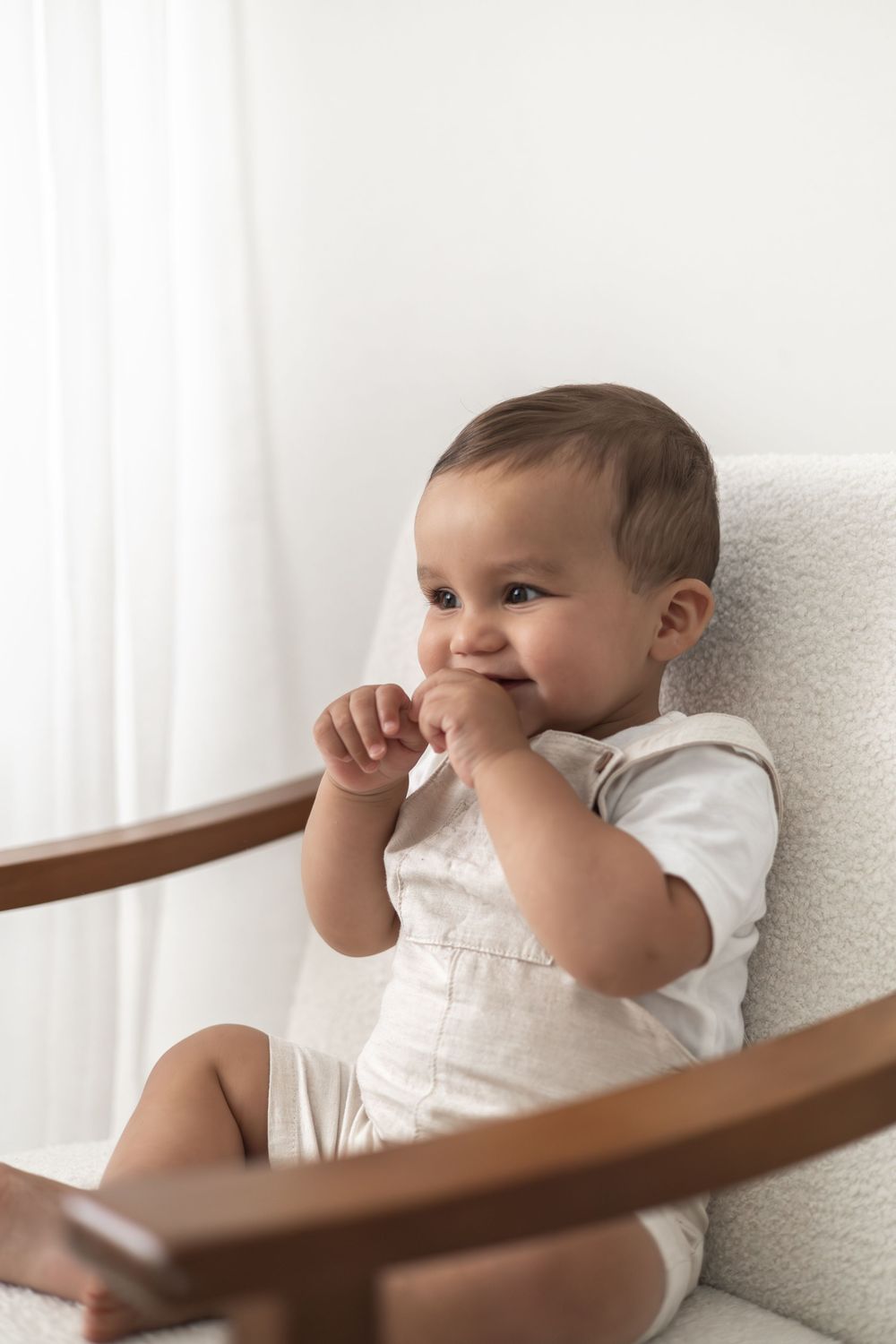 Adorable baby in beige romper sitting in a white chair, smiling Nottingham birthday photoshoot