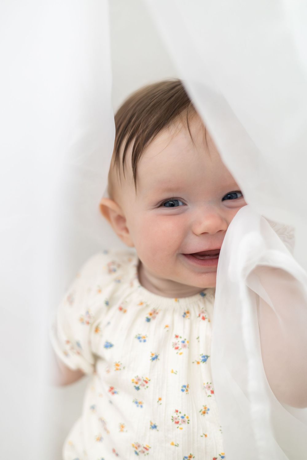 Smiling baby in floral onesie peeking through white curtains, looking happy and playful Birthday photographer Nottingham
