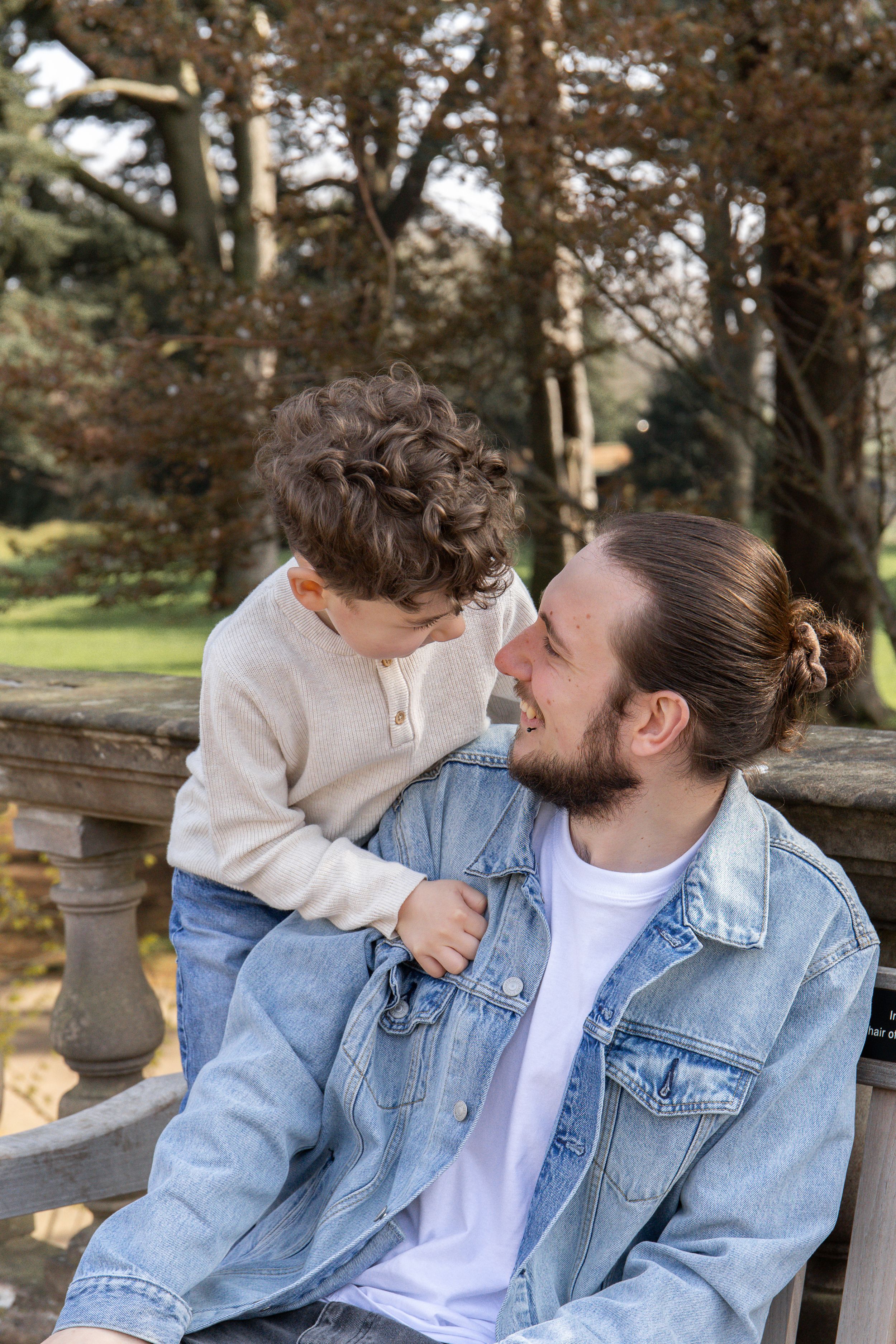 Outdoor family photographer Nottingham A smiling dad in a denim jacket sits on a bench with his son in a park.