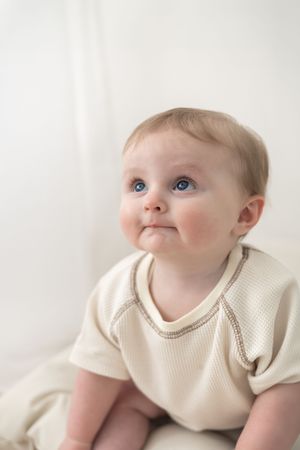 Adorable blue-eyed baby in cream outfit sitting up and looking upward with a curious expression milestone photoshoot Nottingham photographer