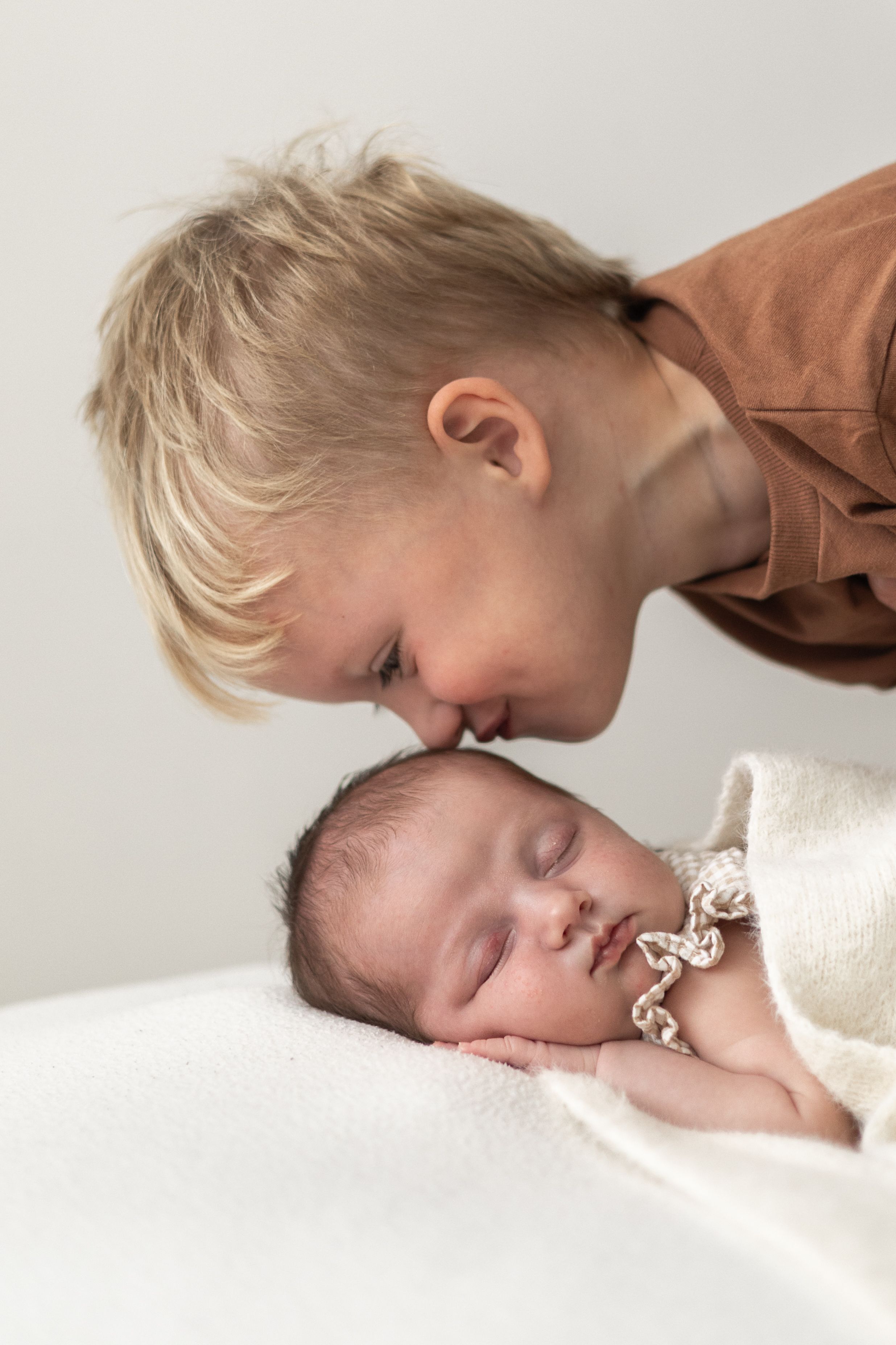Little brother gently kissing sleeping newborn baby wrapped in white blanket Nottingham newborn photography portrait.