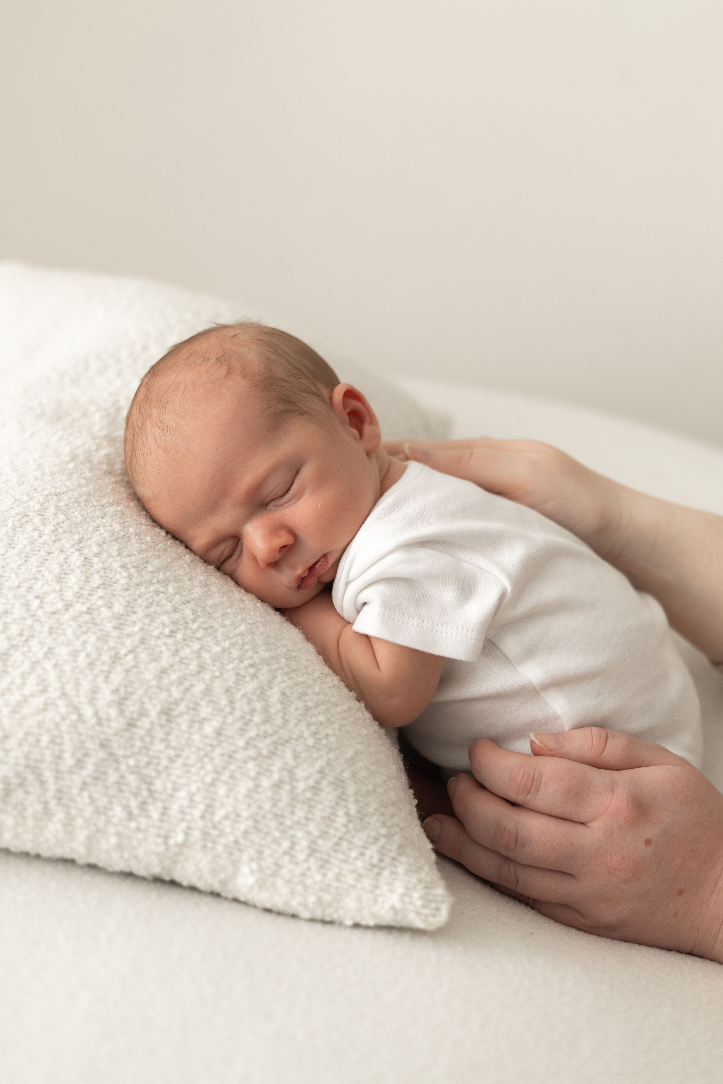Peaceful newborn baby sleeping on white pillow in white onesie with parent's hand nearby natural newborn photography Nottingham NG16
