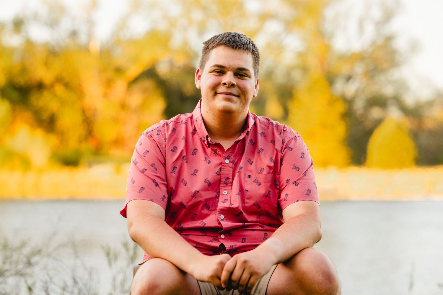 A person in a pink shirt sits outdoors against a vibrant fall foliage background.