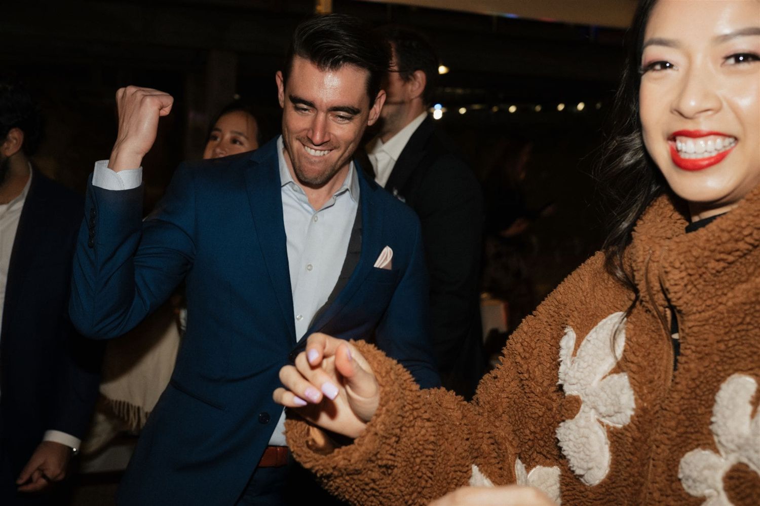 People dancing and celebrating at a joyful evening party, one wearing a blue suit and another in a brown floral sweater.