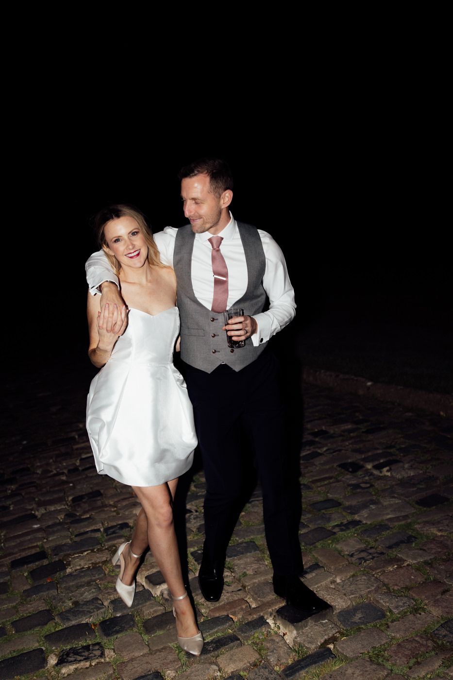 A couple dances playfully on a cobblestone path at night, with the bride in a short white dress.