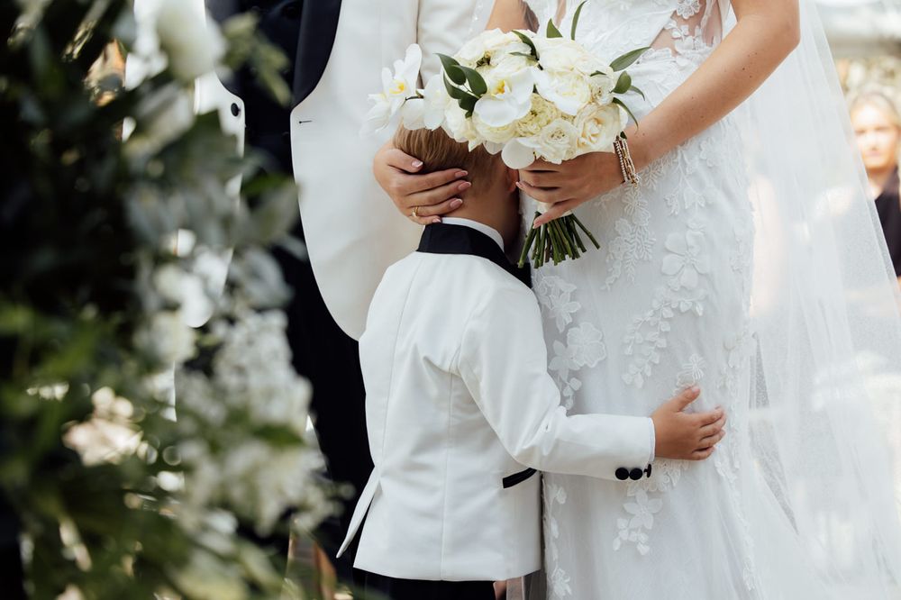 A couple in white wedding attire hold hands during an outdoor ceremony with floral bouquets and natural decor.