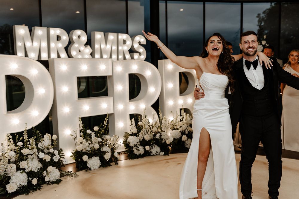 Newlyweds celebrate in front of illuminated MR & MRS marquee letters decorated with white floral arrangements at reception.