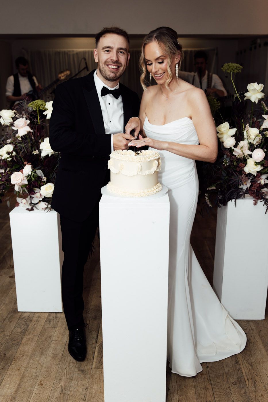 A couple in wedding attire cuts into their elegant white wedding cake displayed on a tall white pedestal surrounded by flowers.