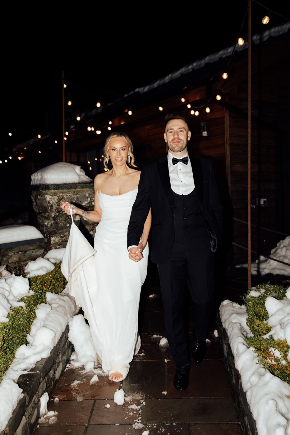 A couple in formal attire walks through snow at night, with the person in a white dress and another in a black tuxedo.
