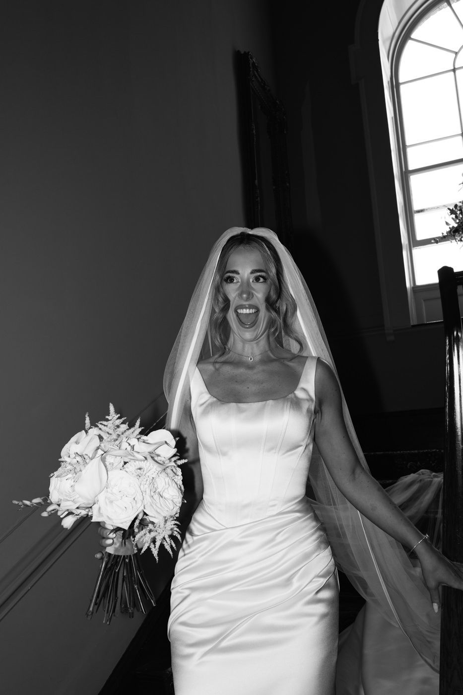 A bride in a sleek white satin dress and veil makes an excited expression while holding a bouquet at peterstone court.