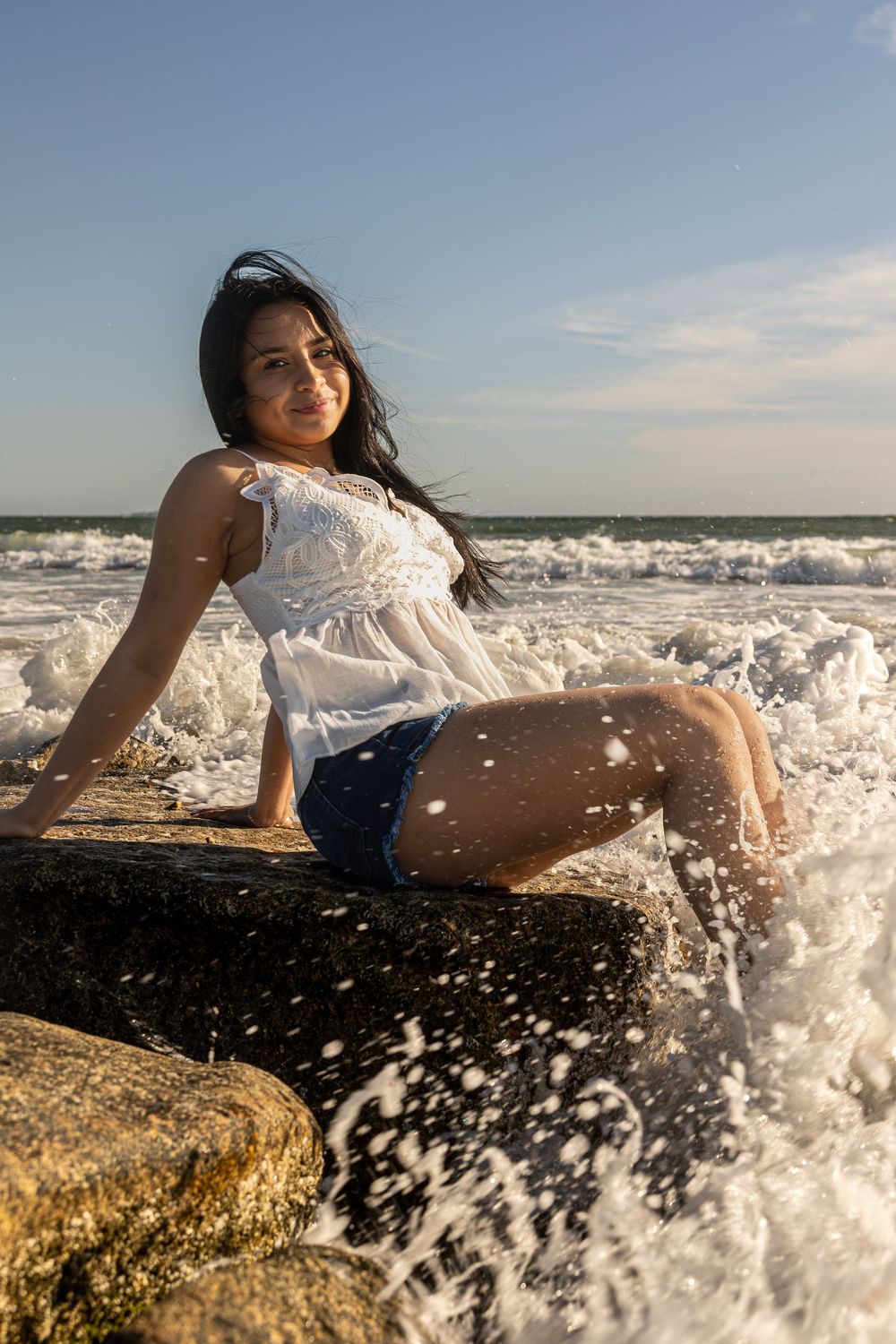 Waves crashing over rocks at beach with white dress against blue sky.