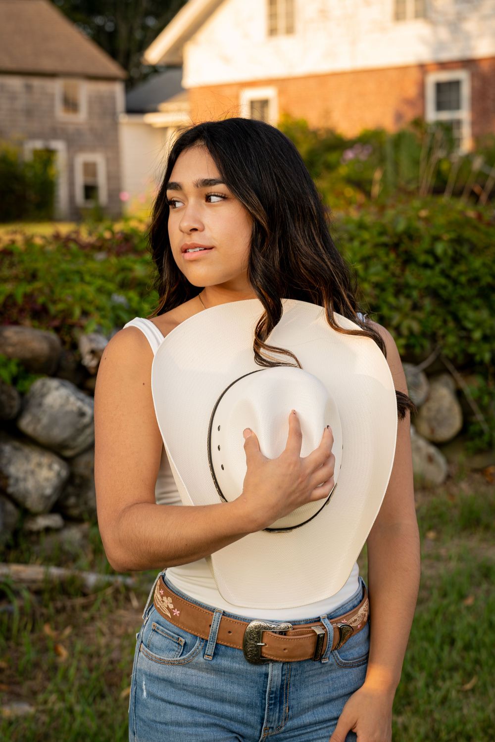 Person holding a white cowboy hat while posing against a rustic house backdrop.