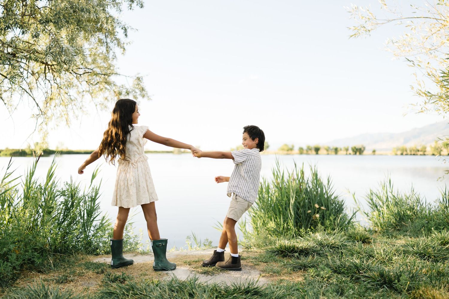 Two children hold hands while playing at the edge of a peaceful lake surrounded by tall grass and trees on a sunny day.