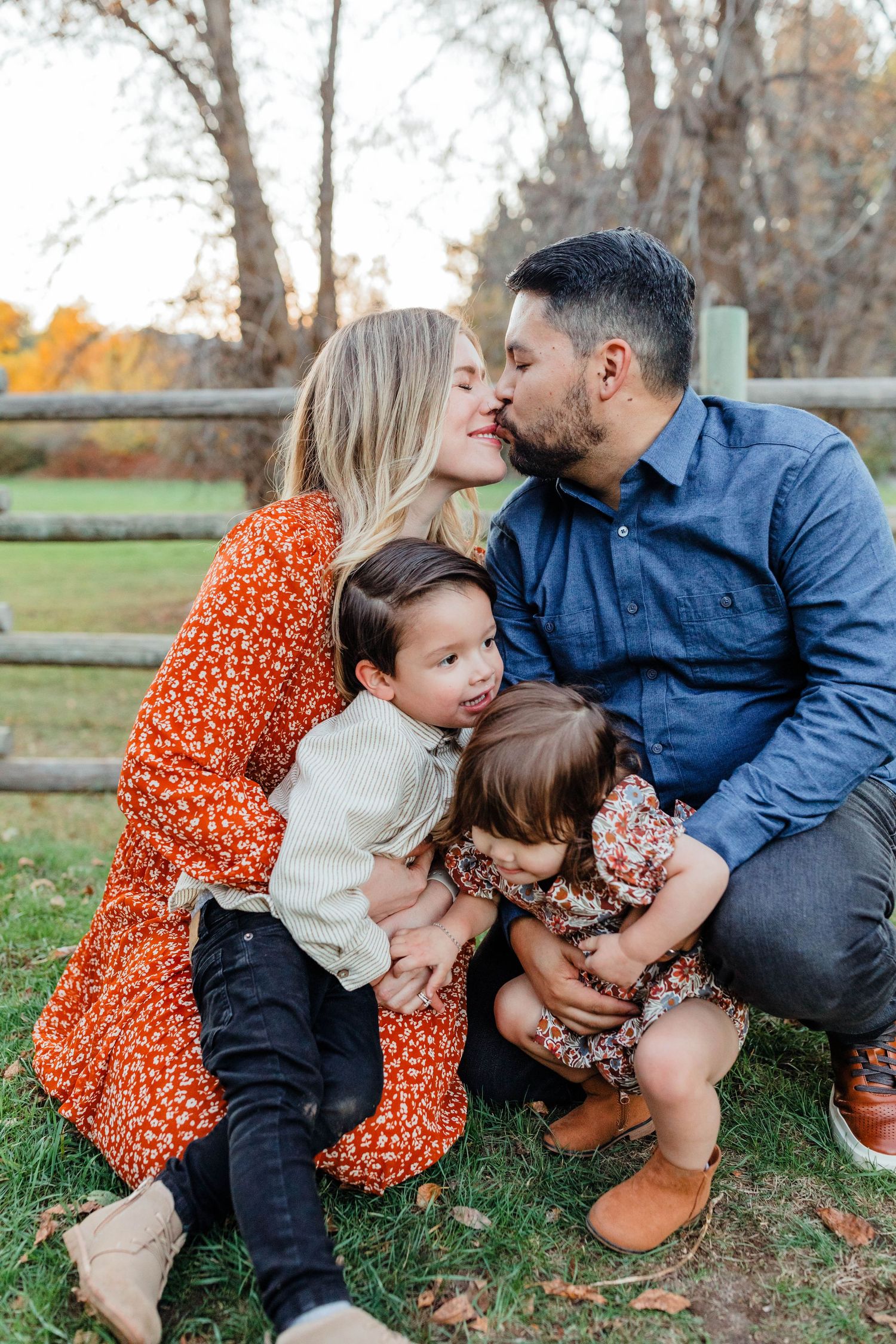 Family of four sitting by wooden fence sharing intimate moment during autumn photoshoot.