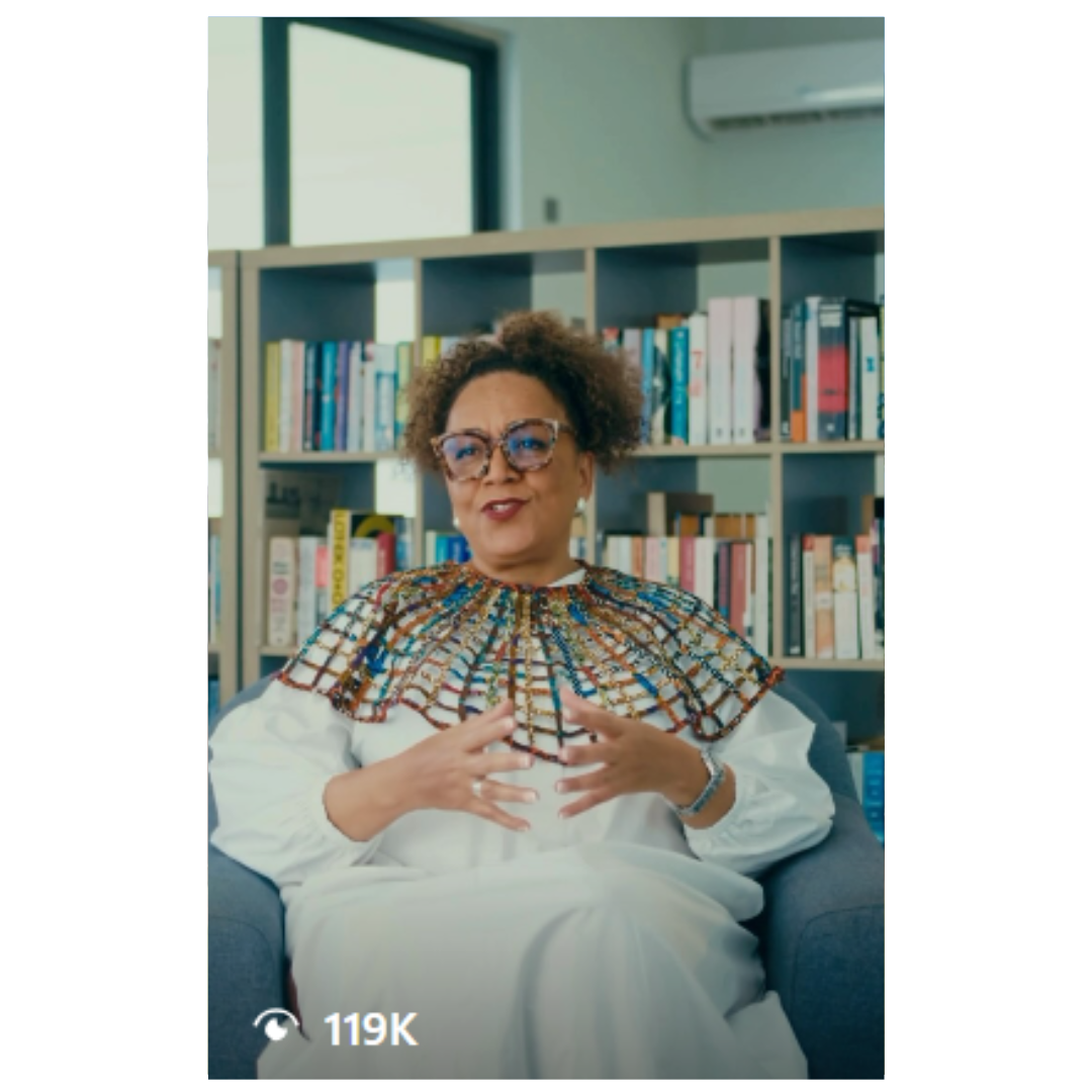 Person seated in library setting with patterned top and white pants against bookshelf background.
