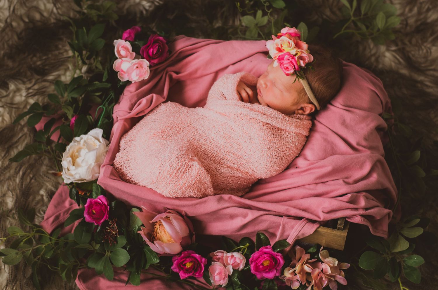 Peaceful newborn swaddled in pink surrounded by pink and white floral arrangements.