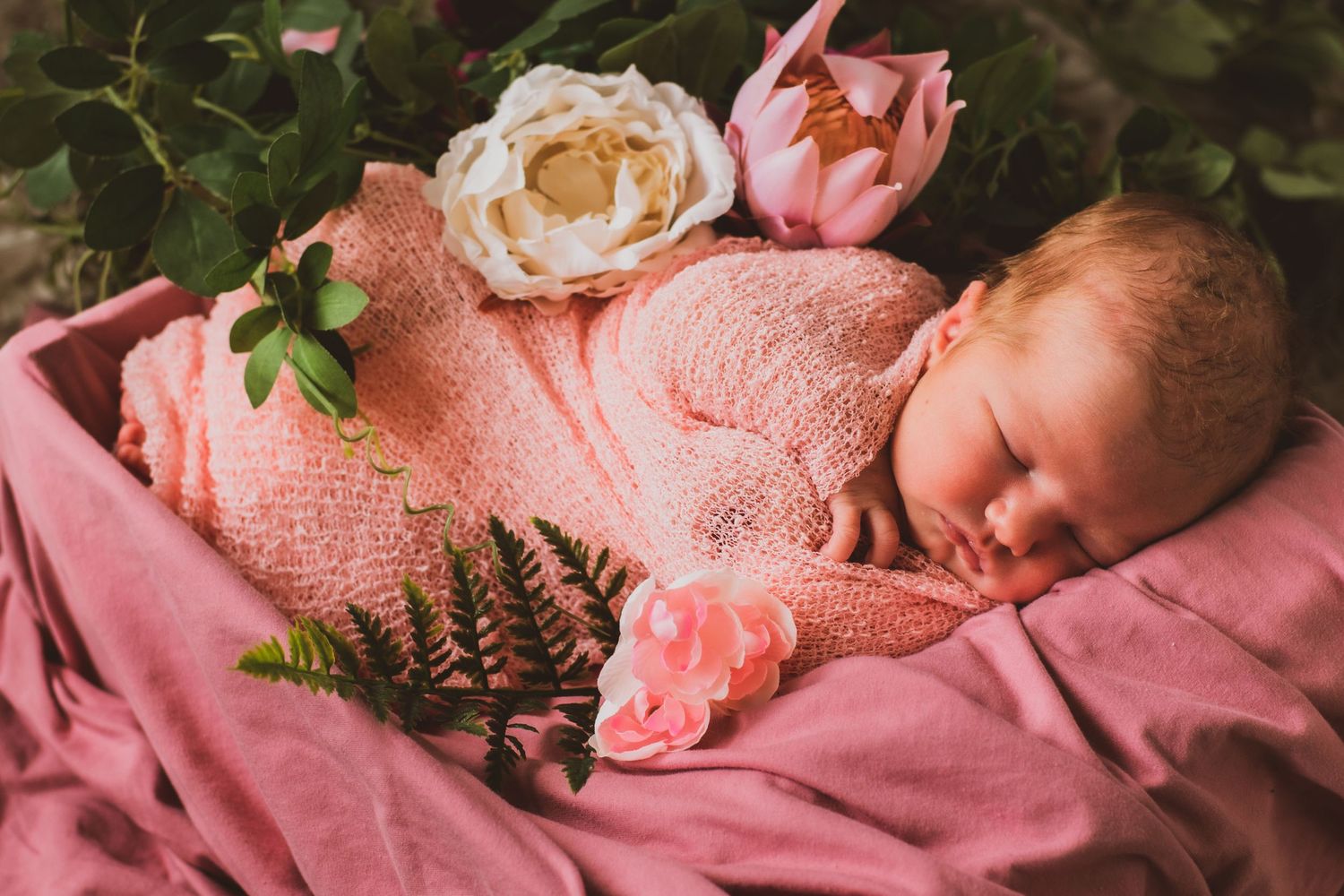 Sleeping newborn wrapped in pink blanket with roses and ferns as decoration.