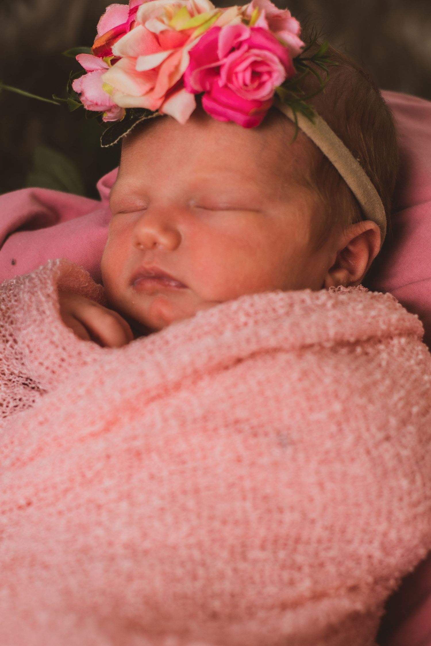 Sleeping newborn wearing floral headband wrapped in pink textured blanket.