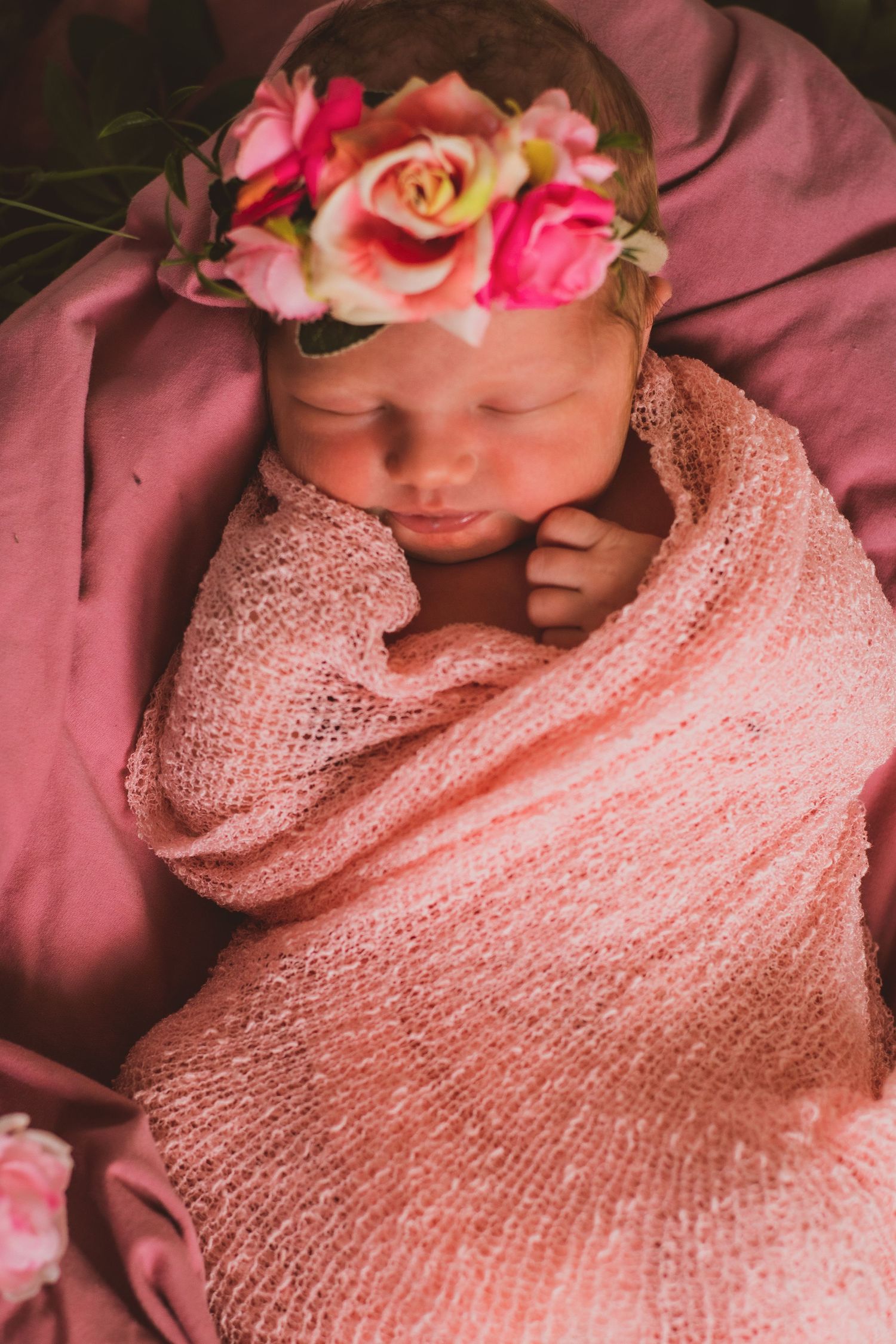 Newborn wrapped in soft pink knit blanket wearing delicate floral crown with pink and peach roses.