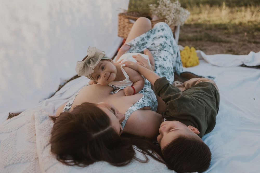 Woman lying on a blanket with a smiling baby on her chest and a young child resting beside them.