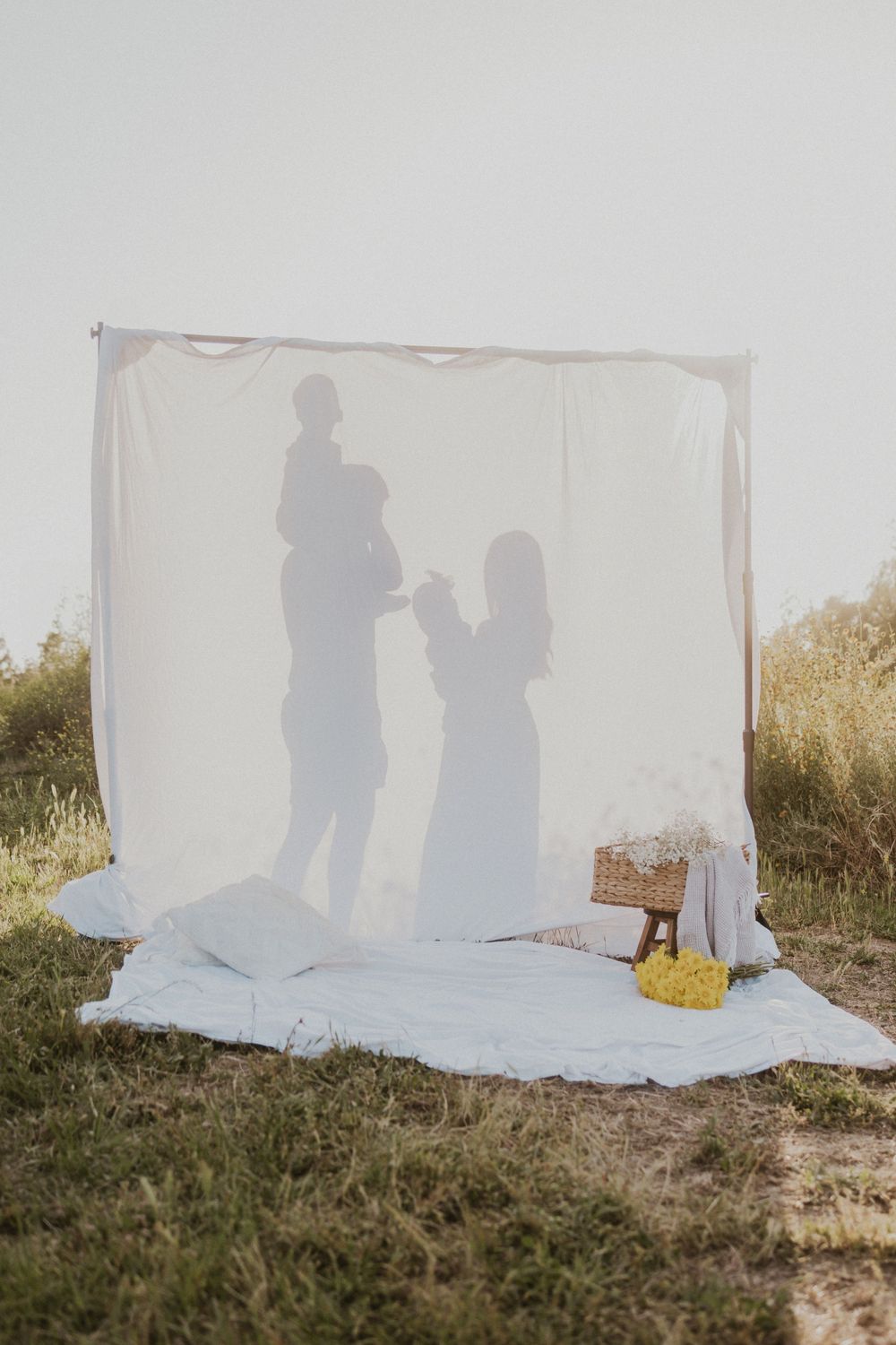 Silhouette of a family with a child on the father's shoulder, in front of a white cloth backdrop in a grassy field.
