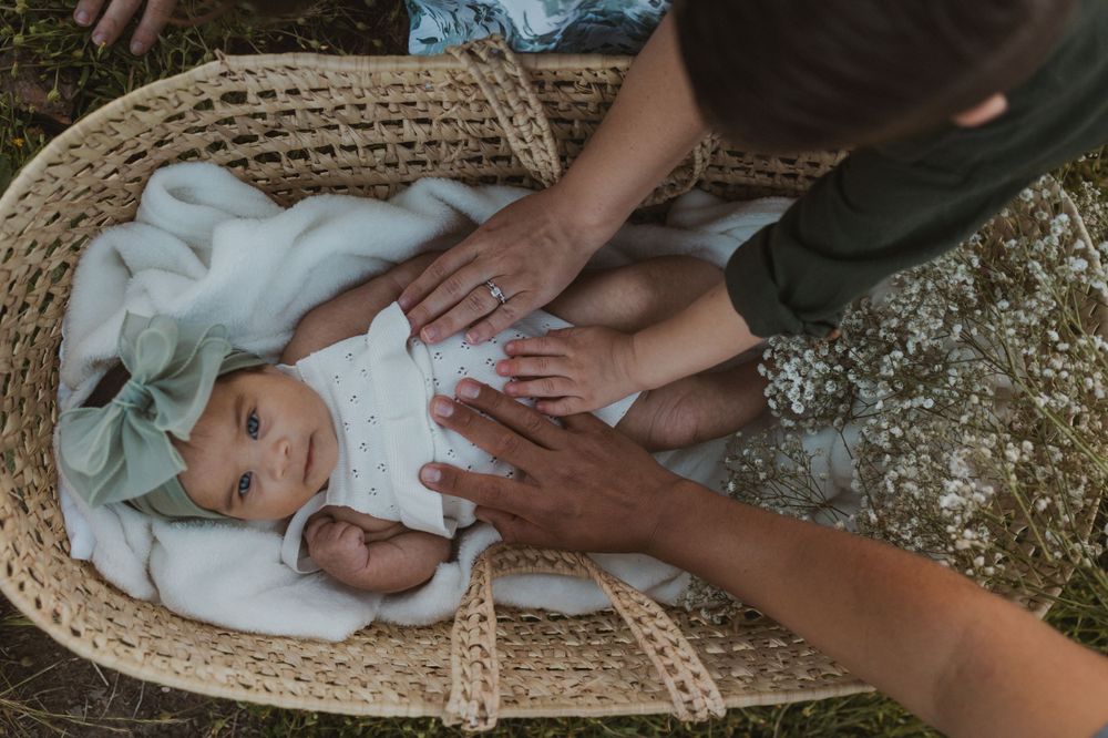 A baby in a wicker basket surrounded by hands, lying on a white blanket, wearing a white outfit & a green bow headband