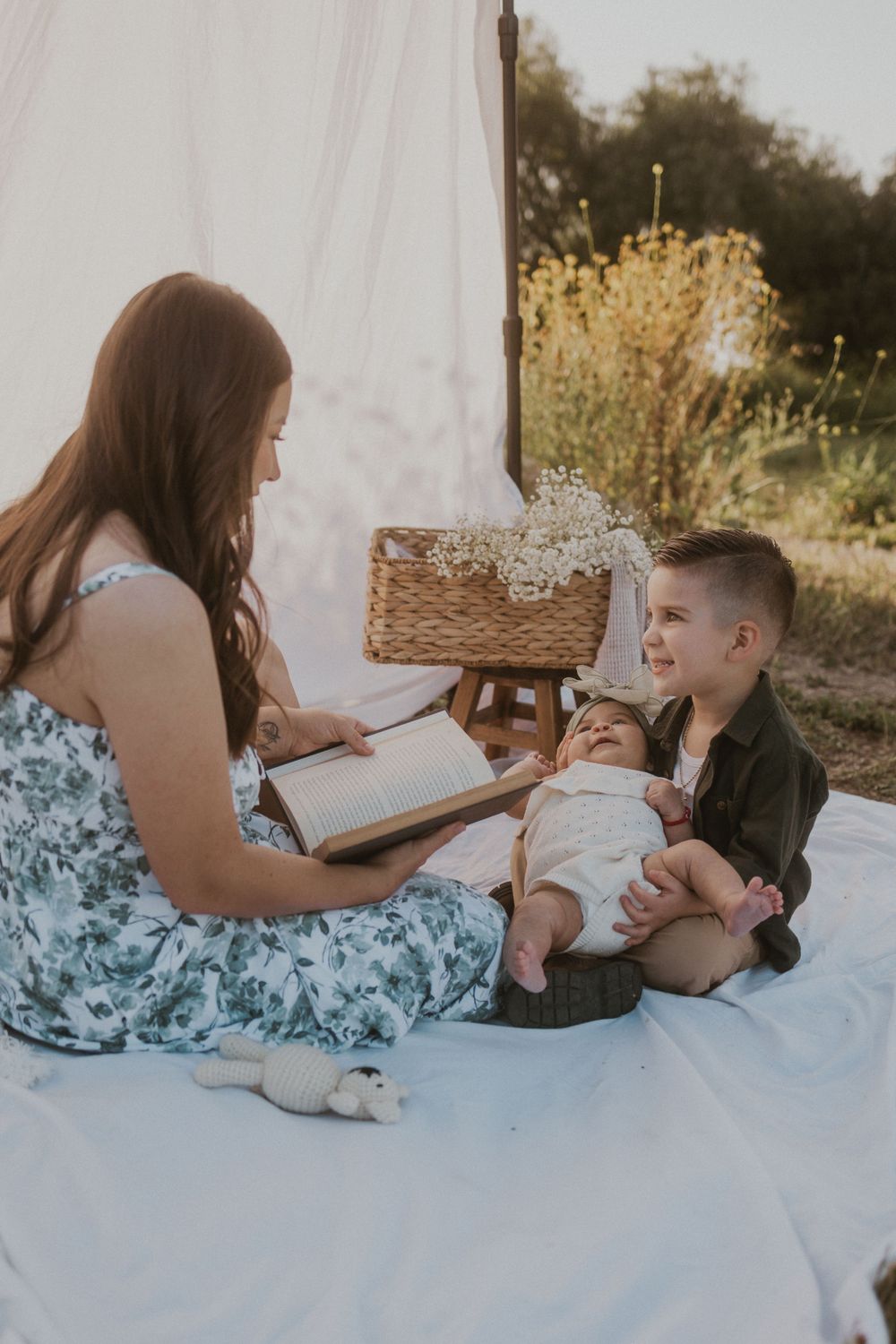 A woman reads a book to a young boy holding a baby, seated on a white blanket outdoors with a basket of flowers